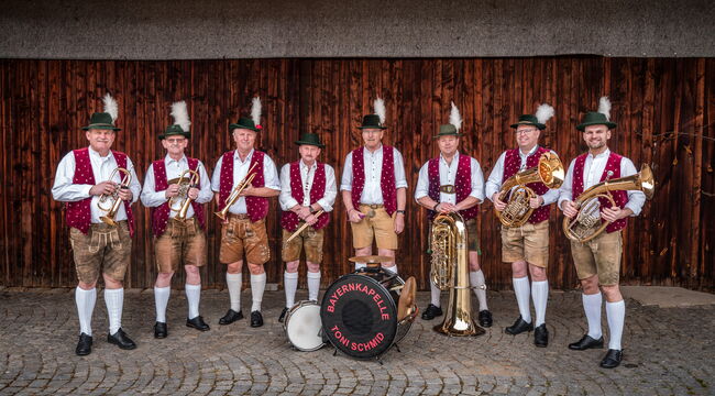 Acht Musiker der Bayernkapelle mit ihren Instrumenten vor einer dunklen Bretterwand