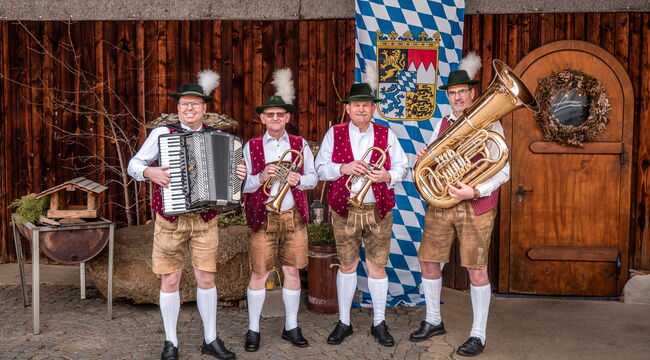 Vier Musiker mit Instrumenten vor einer dunklen Bretterwand mit bayerischer Dekoration